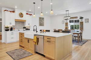 Kitchen with light wood-type flooring, a center island with sink, decorative light fixtures, stainless steel dishwasher, and white cabinetry