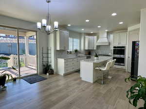 Kitchen with custom exhaust hood, sink, white cabinetry, double oven, and a center island