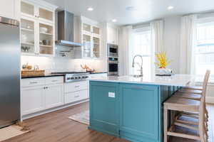 Kitchen with white cabinets, wall chimney exhaust hood, blue cabinetry, stainless steel appliances, and light hardwood / wood-style floors