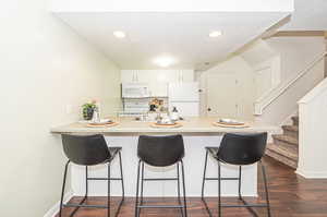 Kitchen featuring white appliances, dark hardwood / wood-style flooring, kitchen peninsula, white cabinetry, and a kitchen bar