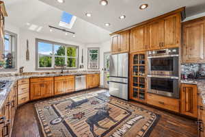 Kitchen with vaulted ceiling with skylight, light stone counters, recessed lighting, stainless steel appliances, and dark wood-style floors