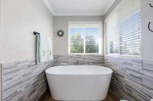 Bathroom with crown molding, a tub to relax in, and tile walls