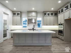 Kitchen featuring a center island with sink, gray cabinetry, double oven, and extractor fan