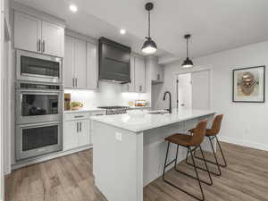 Kitchen featuring an island with sink, custom range hood, stainless steel appliances, sink, and light hardwood / wood-style floors