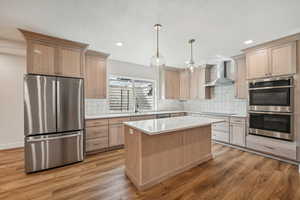 Kitchen featuring light wood-type flooring, a kitchen island, hanging light fixtures, stainless steel appliances, and wall chimney exhaust hood