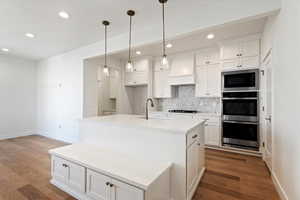 Kitchen with wood flooring, an island with built in bench, white cabinets, double ovens and custom range hood