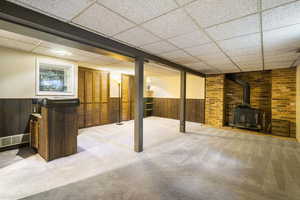 Basement featuring a paneled ceiling, wood walls, carpet, and a wood stove