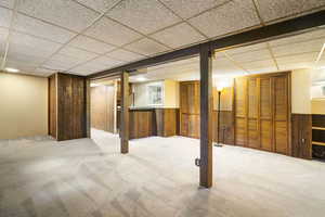 Basement with carpet flooring, a drop ceiling, and wooden walls