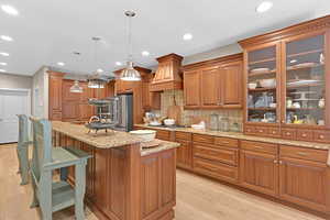 Kitchen with decorative backsplash, a center island with sink, decorative light fixtures, custom exhaust hood, and light hardwood / wood-style floors