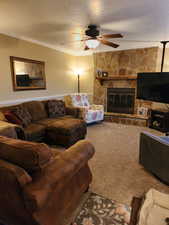 Carpeted living room featuring a stone fireplace, ornamental molding, a textured ceiling, and ceiling fan