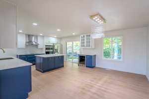 Kitchen with wall chimney range hood, stainless steel appliances, blue cabinetry, white cabinetry, and light hardwood / wood-style floors