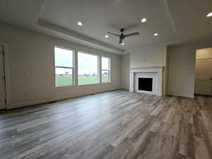 Unfurnished living room featuring a fireplace, a tray ceiling, light wood-type flooring, and ceiling fan