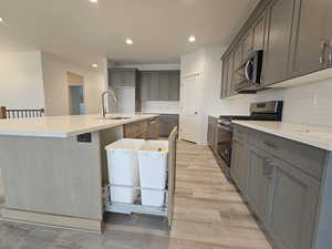 Kitchen with appliances with stainless steel finishes, gray cabinetry, a center island with sink, and light hardwood / wood-style floors