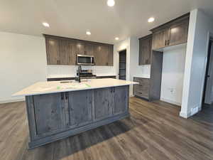 Kitchen featuring appliances with stainless steel finishes, dark brown cabinets, dark wood-type flooring, decorative backsplash, and a center island with sink