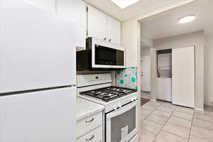 Kitchen featuring white appliances, light tile patterned flooring, backsplash, stacked washer and dryer, and white cabinets