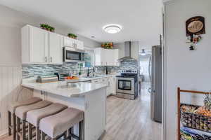 Kitchen featuring a breakfast bar area, appliances with stainless steel finishes, wall chimney range hood, and white cabinets