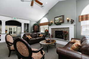 Carpeted living area featuring high vaulted ceiling, a ceiling fan, decorative columns, and a fireplace with raised hearth