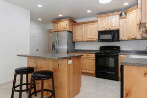 Kitchen with black appliances, light brown cabinets, dark countertops, a sink, and a kitchen island