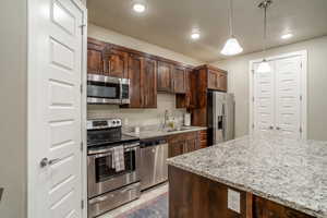 Kitchen featuring light stone countertops, appliances with stainless steel finishes, dark brown cabinets, and hanging light fixtures