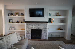 Living room with a tiled fireplace, dark wood-type flooring, and built in features