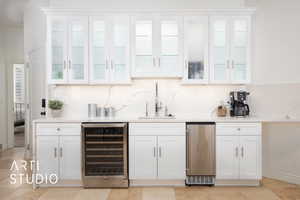 Bar featuring wine cooler, sink, white illuminated cabinets, and backsplash