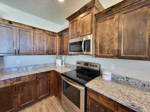 Kitchen featuring dark brown cabinetry, light stone countertops, stainless steel appliances, and light wood-type flooring