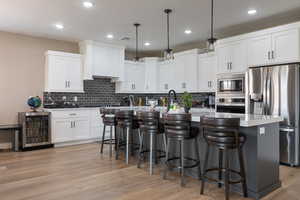 Kitchen with appliances with stainless steel finishes, hanging light fixtures, wine cooler, light hardwood / wood-style flooring, and a kitchen island with sink