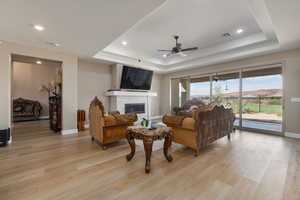 Living room featuring light hardwood / wood-style floors, a stone fireplace, a raised ceiling, and ceiling fan