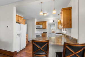 Kitchen featuring dark wood-type flooring, hanging light fixtures, kitchen peninsula, a kitchen bar, and white appliances