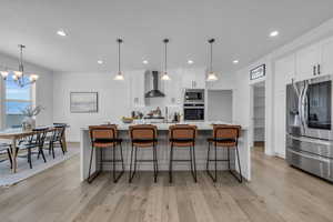 Kitchen with light wood-type flooring, stainless steel appliances, wall chimney exhaust hood, white cabinets, and a center island with sink