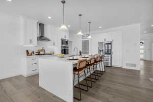 Kitchen featuring wall chimney range hood, an island with sink, appliances with stainless steel finishes, white cabinetry, and light hardwood / wood-style flooring