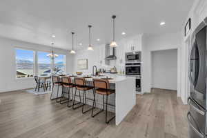 Kitchen with wall chimney range hood, appliances with stainless steel finishes, an island with sink, white cabinetry, and pendant lighting