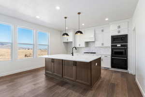 Kitchen featuring white cabinets, custom range hood, a center island with sink, and dark wood-type flooring