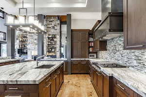 Kitchen featuring light stone countertops, light wood-type flooring, wall chimney exhaust hood, sink, and a large island with sink