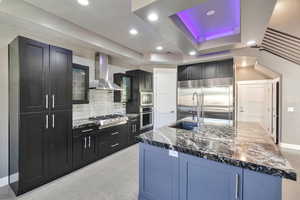 Kitchen with a large island, tasteful backsplash, a tray ceiling, built in appliances, and wall chimney exhaust hood