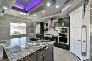 Kitchen with tasteful backsplash, appliances with stainless steel finishes, a raised ceiling, wall chimney exhaust hood, and a kitchen island with sink