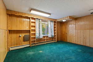 Bedroom with wood walls, a textured ceiling, a baseboard radiator, and dark colored carpet  This room has an outside entrance and has been used in the past as an office for work.