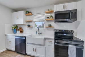 Kitchen with tasteful backsplash, appliances with black stainless steel finishes, farmhouse sink, light wood-type flooring, and white cabinetry with open shelving