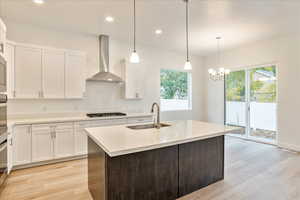 Kitchen with wall chimney range hood, stainless steel gas stovetop, a kitchen island with sink, light wood-type flooring, and sink