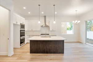 Kitchen featuring stainless steel appliances, wall chimney exhaust hood, white cabinets, light hardwood / wood-style flooring, and a center island with sink