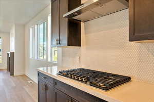 Kitchen with tasteful backsplash, light wood-type flooring, stainless steel gas cooktop, dark brown cabinets, and wall chimney exhaust hood