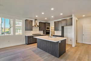 Kitchen with wall chimney range hood, an island with sink, sink, appliances with stainless steel finishes, and light hardwood / wood-style floors