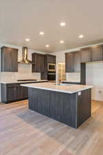 Kitchen featuring wall chimney range hood, a center island with sink, sink, appliances with stainless steel finishes, and light hardwood / wood-style floors