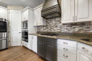 Kitchen with premium range hood, stainless steel appliances, wood-type flooring, dark stone countertops, and white cabinets