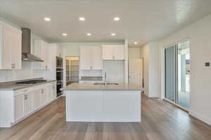 Kitchen with a kitchen island with sink, wall chimney exhaust hood, sink, and white cabinets