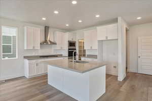 Kitchen with wall chimney range hood, sink, an island with sink, and white cabinets