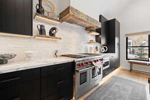 Kitchen with light stone countertops, tasteful backsplash, custom exhaust hood, double oven range, and light hardwood / wood-style floors