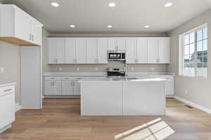 Kitchen with appliances with stainless steel finishes, light hardwood / wood-style flooring, white cabinetry, and a kitchen island with sink