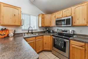 Kitchen featuring lofted ceiling, light tile patterned floors, appliances with stainless steel finishes, light brown cabinetry, and sink