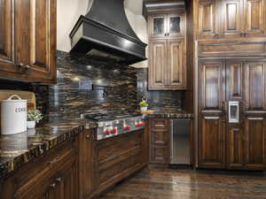 Kitchen with tasteful backsplash, appliances with stainless steel finishes, dark hardwood / wood-style flooring, dark stone counters, and premium range hood